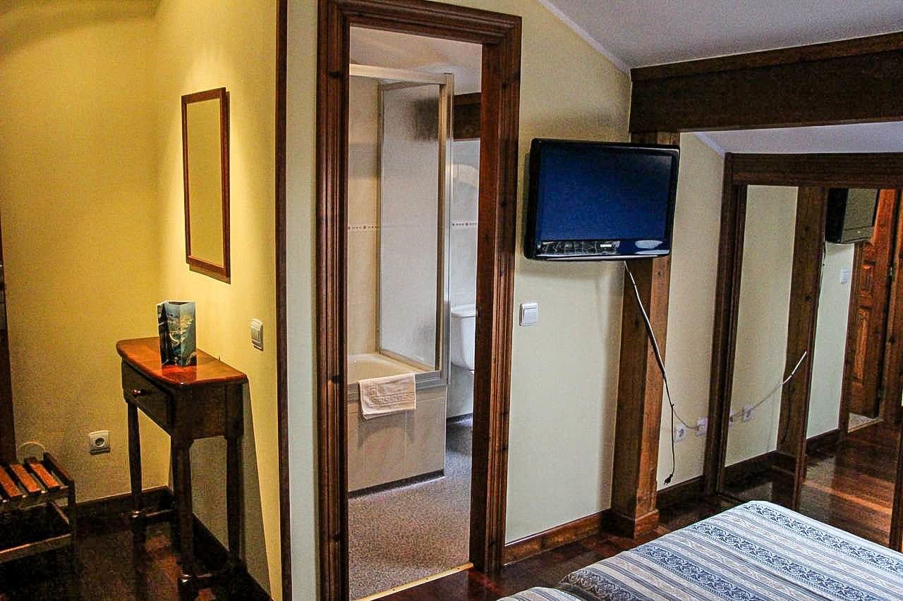 Habitación de hotel con piso de madera, cama con colcha estampada, televisor montado en la pared, mesita de noche de madera con un libro y un espejo en la pared.