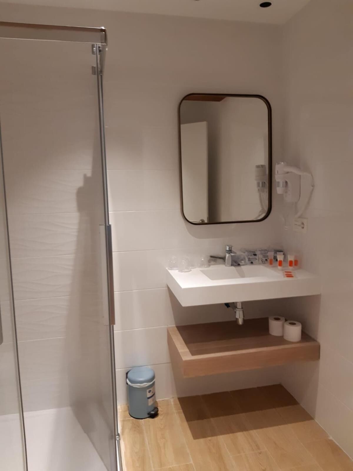 A modern bathroom with a glass shower enclosure, a white sink with a mirror above it, and a wooden shelf below.