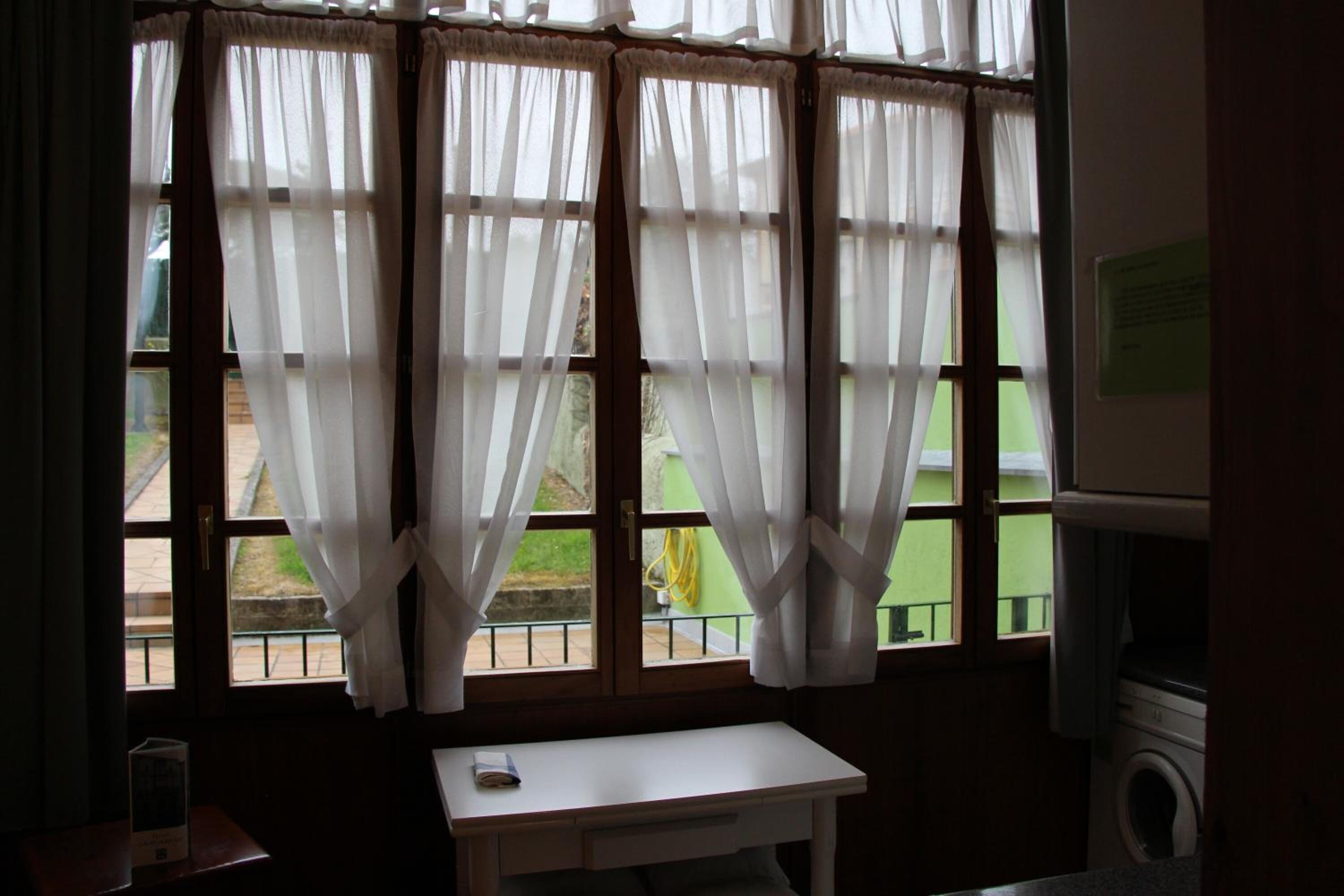 A room with a window featuring white curtains, a small table with a book, and a washing machine in the corner.