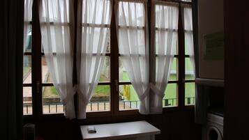 A room with a window featuring white curtains, a small table with a book, and a washing machine in the corner.