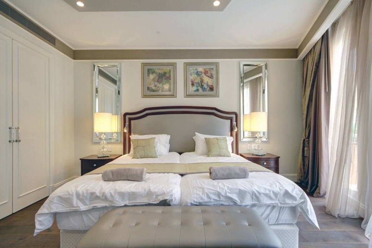 Une chambre spacieuse avec un grand lit, deux tables de chevet avec des lampes, un miroir et des œuvres d’art accrochées au mur.