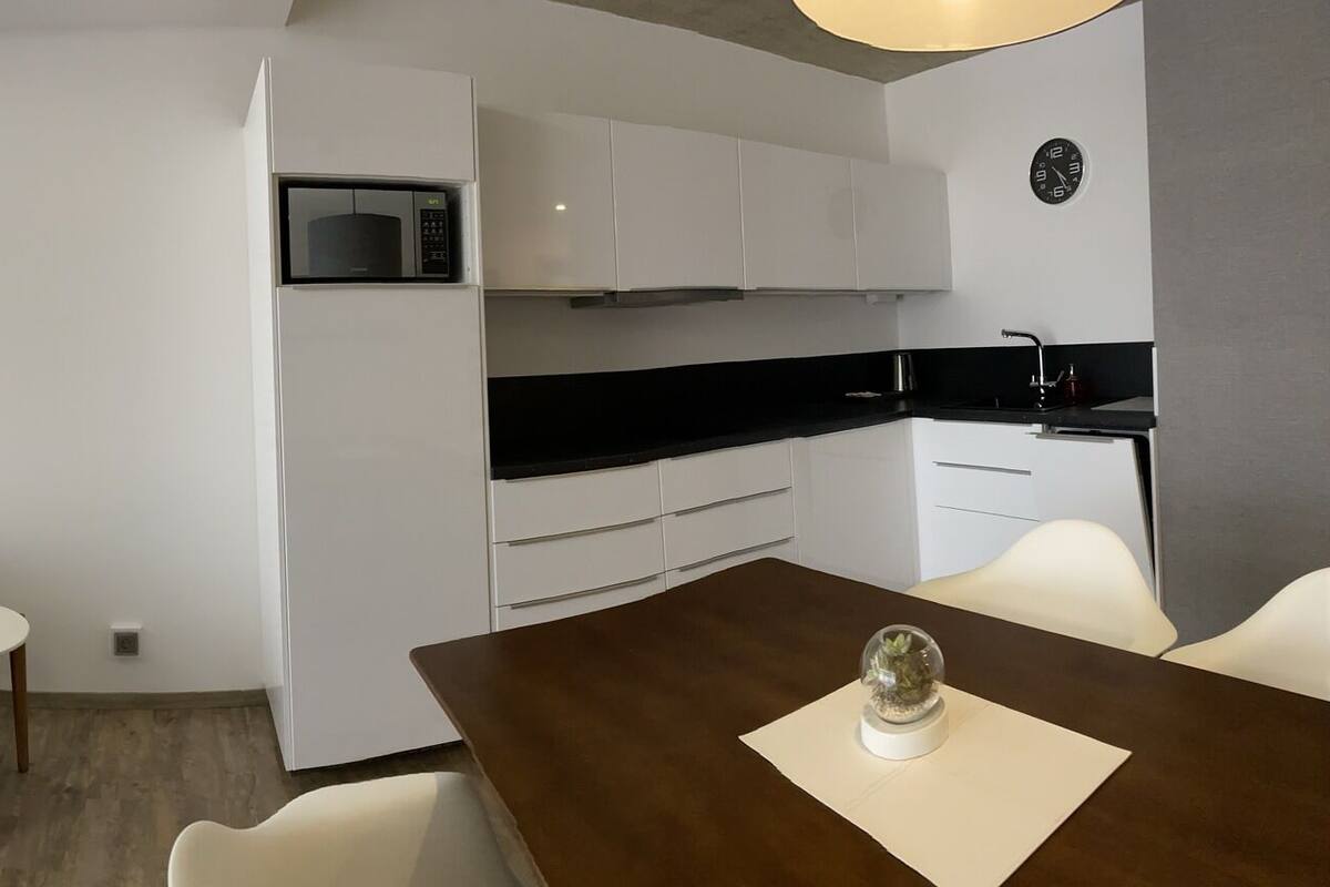 Una habitación de hotel moderna con televisión de pantalla plana, un sofá, una mesa de comedor, una zona de cocina y un amplio ventanal con cortinas translúcidas.