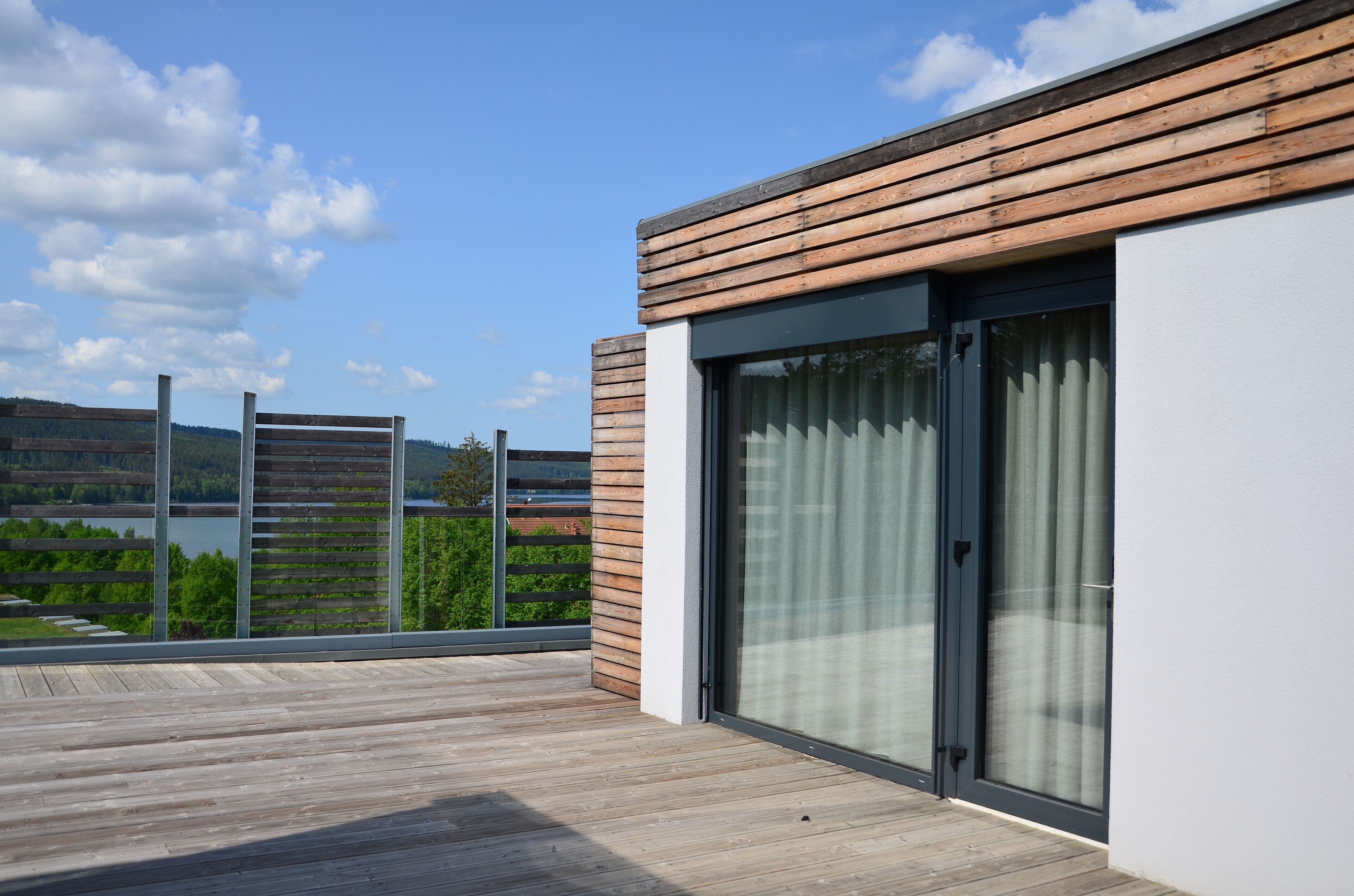 A modern balcony with a wooden floor and a glass door, overlooking a lake and greenery.