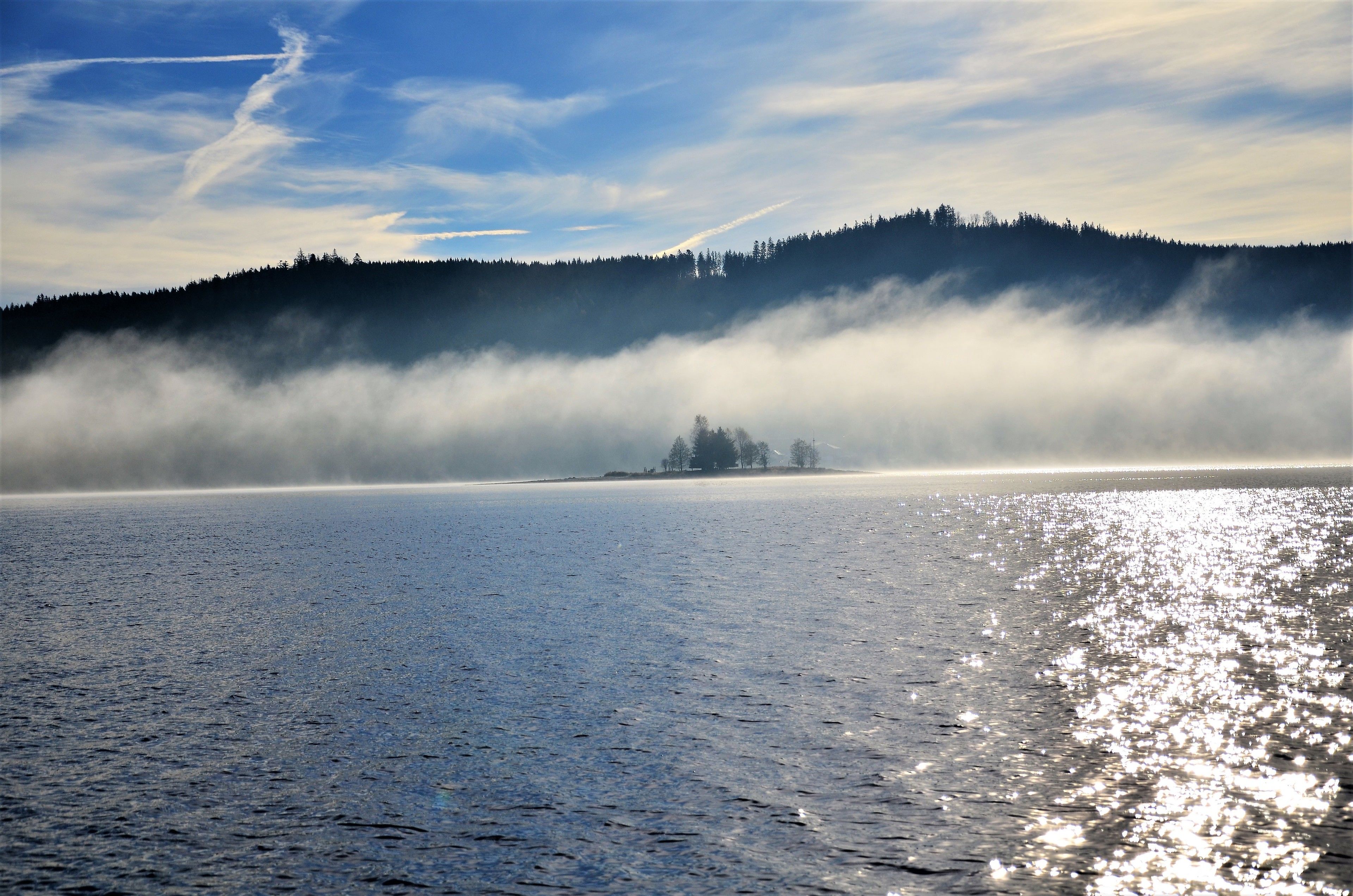 A calm body of water reflecting sunlight, a forested hill in the background, and a misty atmosphere.
