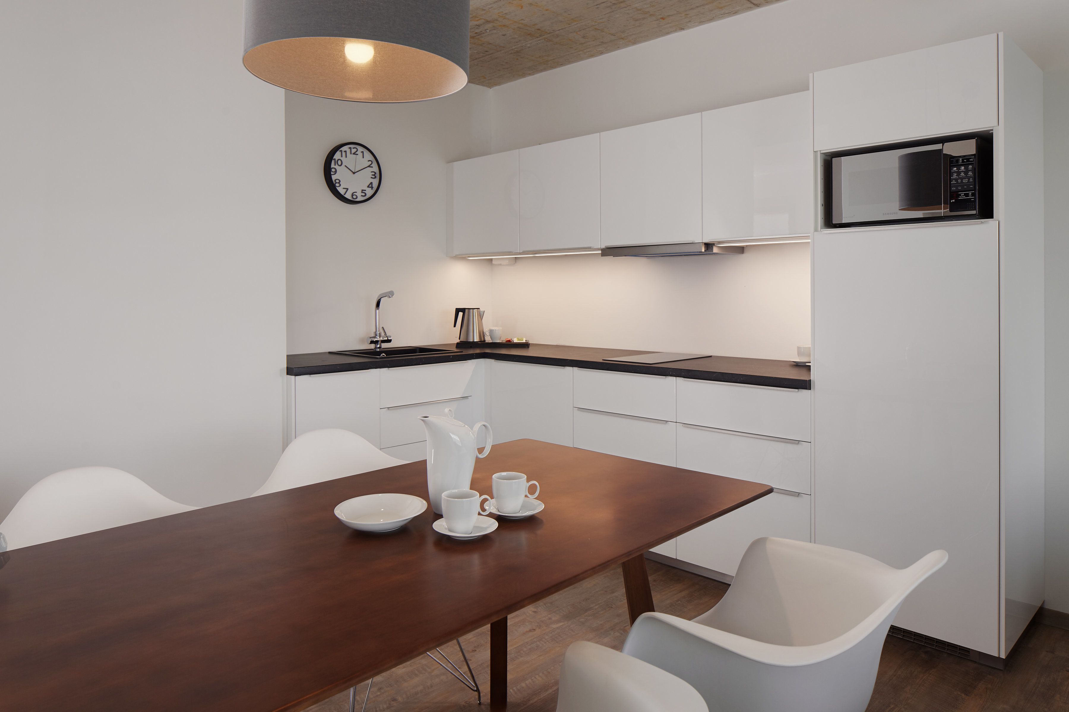 A modern kitchen with white cabinets, a wooden dining table, white chairs, a clock, a microwave, and a coffee maker.