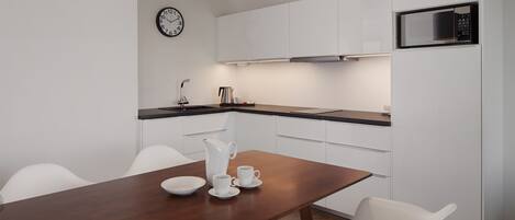 A modern kitchen with white cabinets, a wooden dining table, white chairs, a clock, a microwave, and a coffee maker.