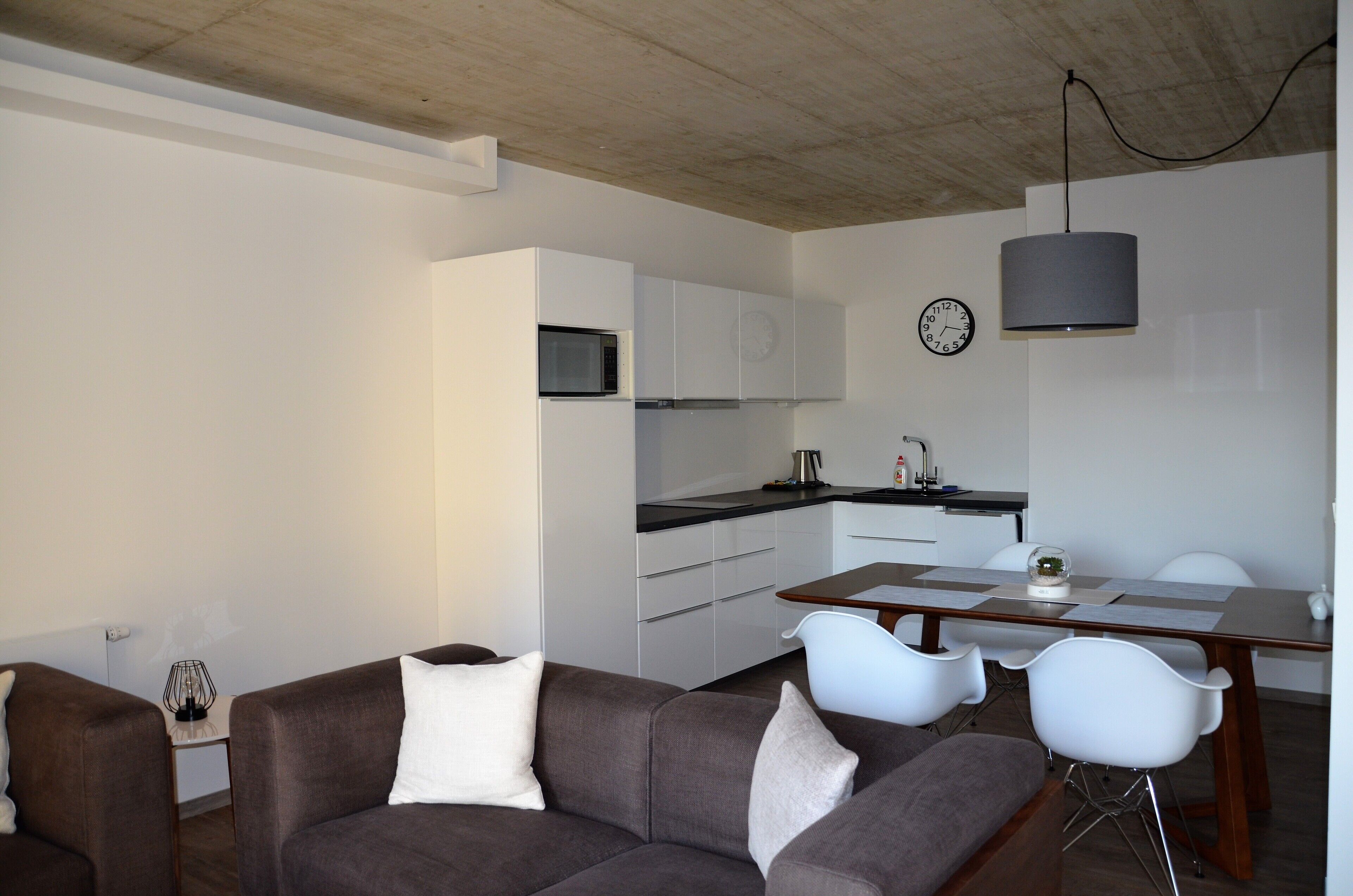 A modern living room with a brown sofa and white chairs, a dining table with a lamp, and a kitchen area with white cabinets and a clock on the wall.