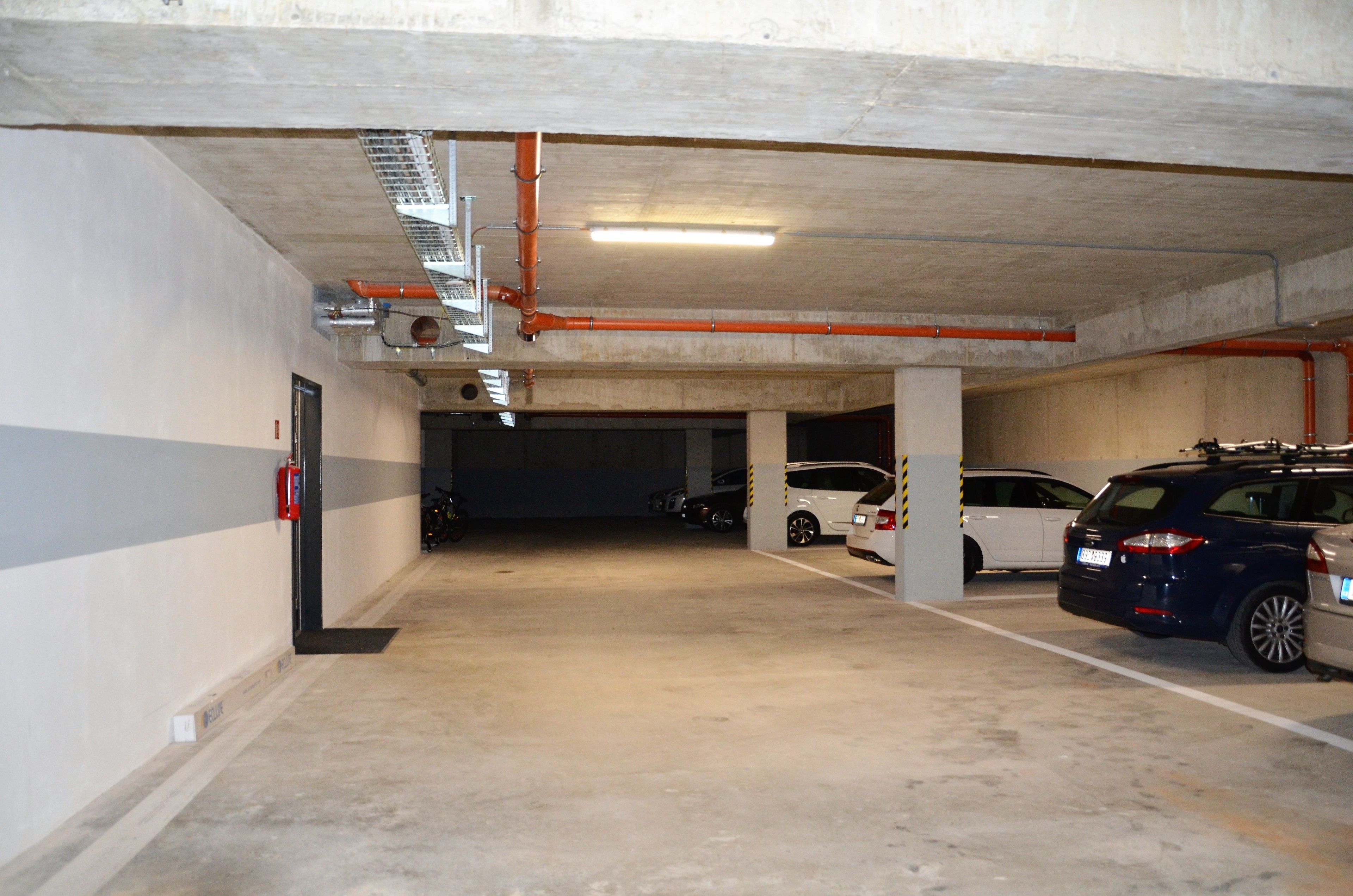 A multi-level parking garage with concrete pillars, fluorescent lighting, and a fire extinguisher on the wall.