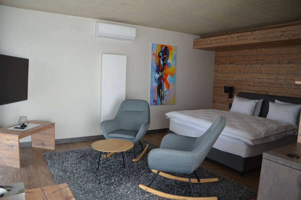 Una habitación de hotel moderna con una cama, dos sillones, una mesita, un televisor y un cuadro en la pared.