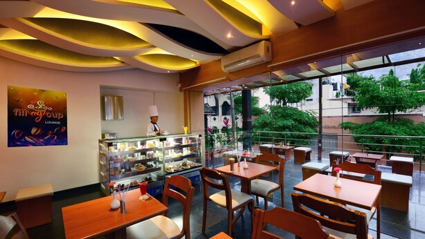 A modern cafe with wooden tables and chairs, a glass display case with desserts, and a view of greenery outside.