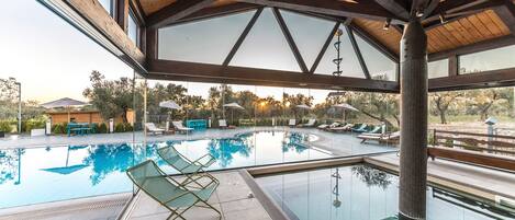 A modern indoor swimming pool with clear glass walls, lounge chairs, and umbrellas.