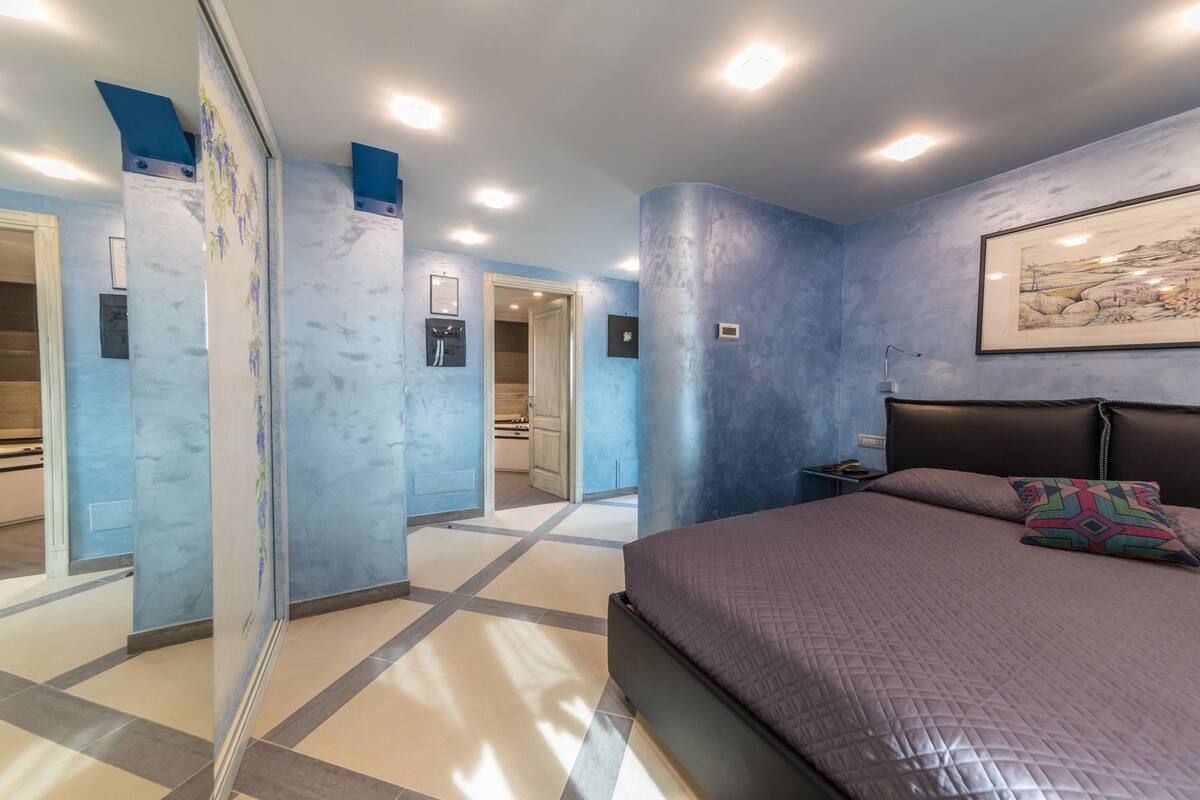 A bedroom with a bed, a nightstand, a framed picture on the wall, and a bathroom visible through a glass door.