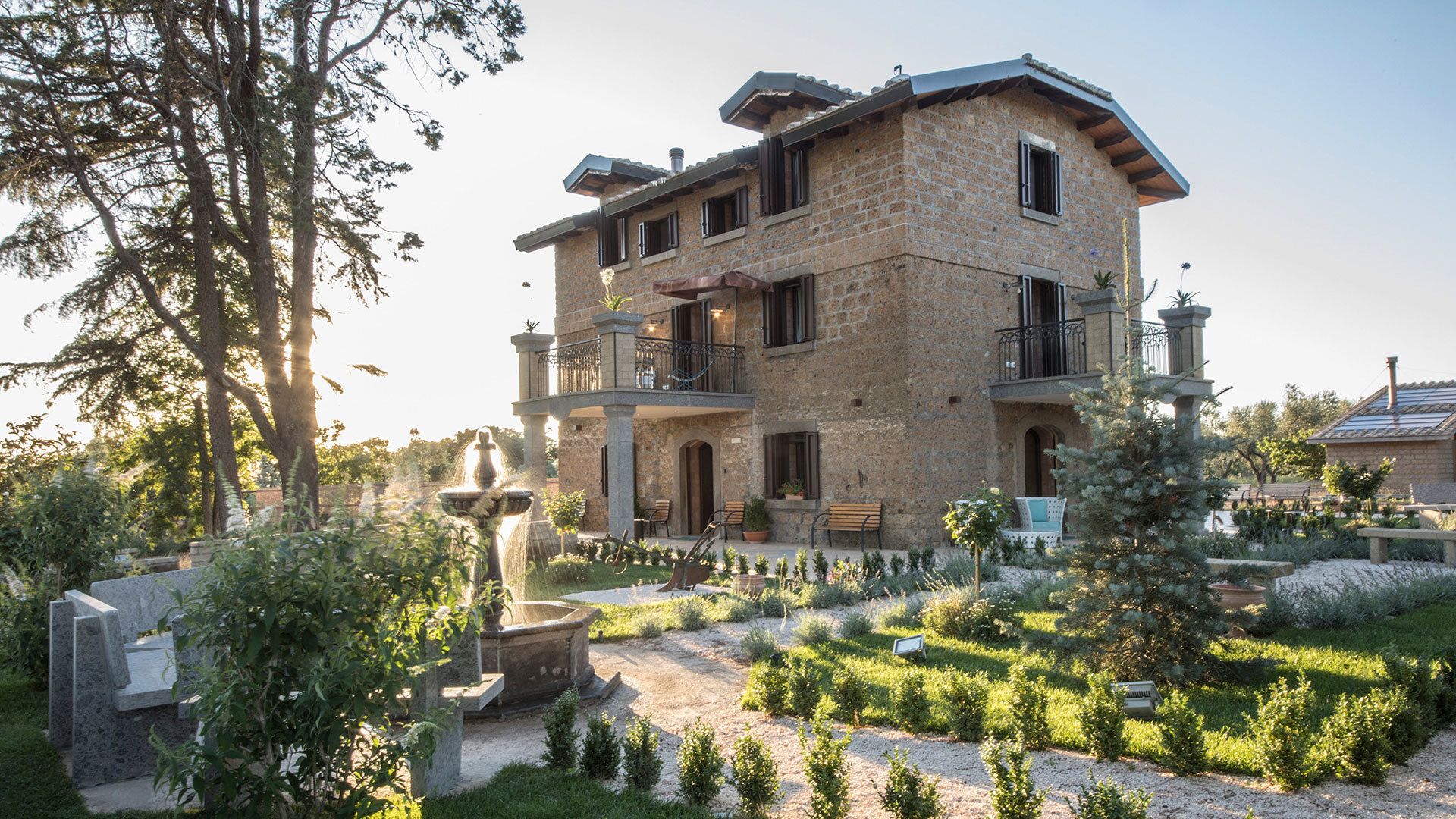 A two-story stone building with a balcony, surrounded by a well-maintained garden with various plants and a fountain.