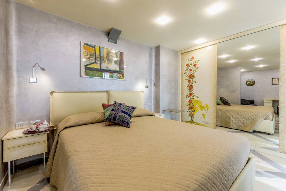 A bedroom with a large bed, a bedside table with a lamp, a mirror, and a painting on the wall.