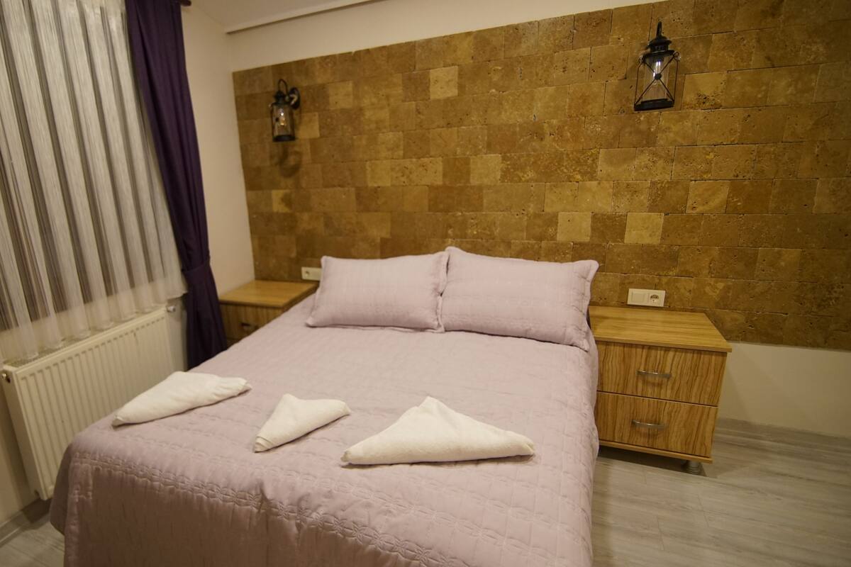 A hotel room with a bed, two nightstands, a wall-mounted lamp, and a window with curtains.