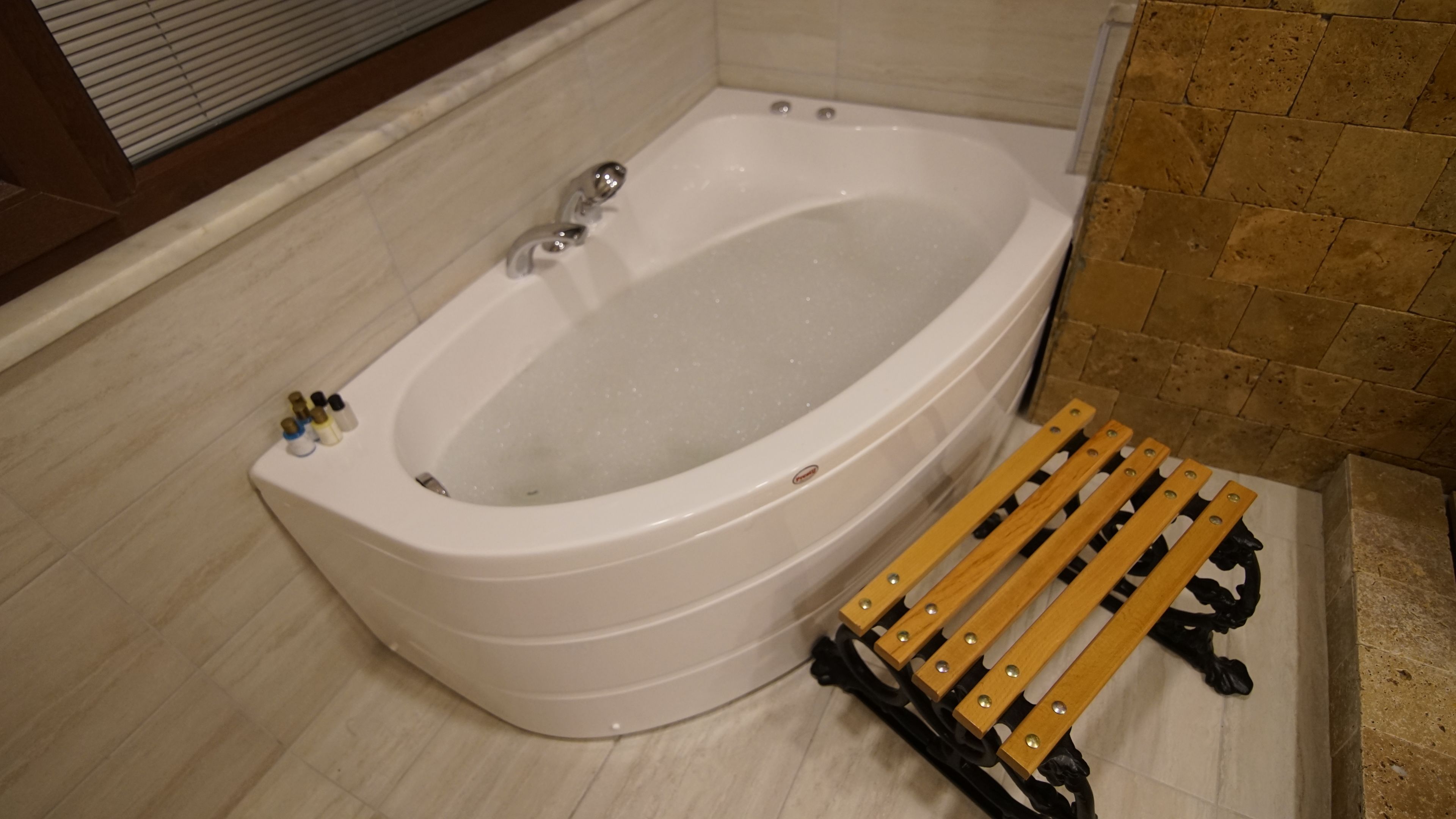 A white bathtub with a wooden bench, a set of toiletries, and a tiled wall.