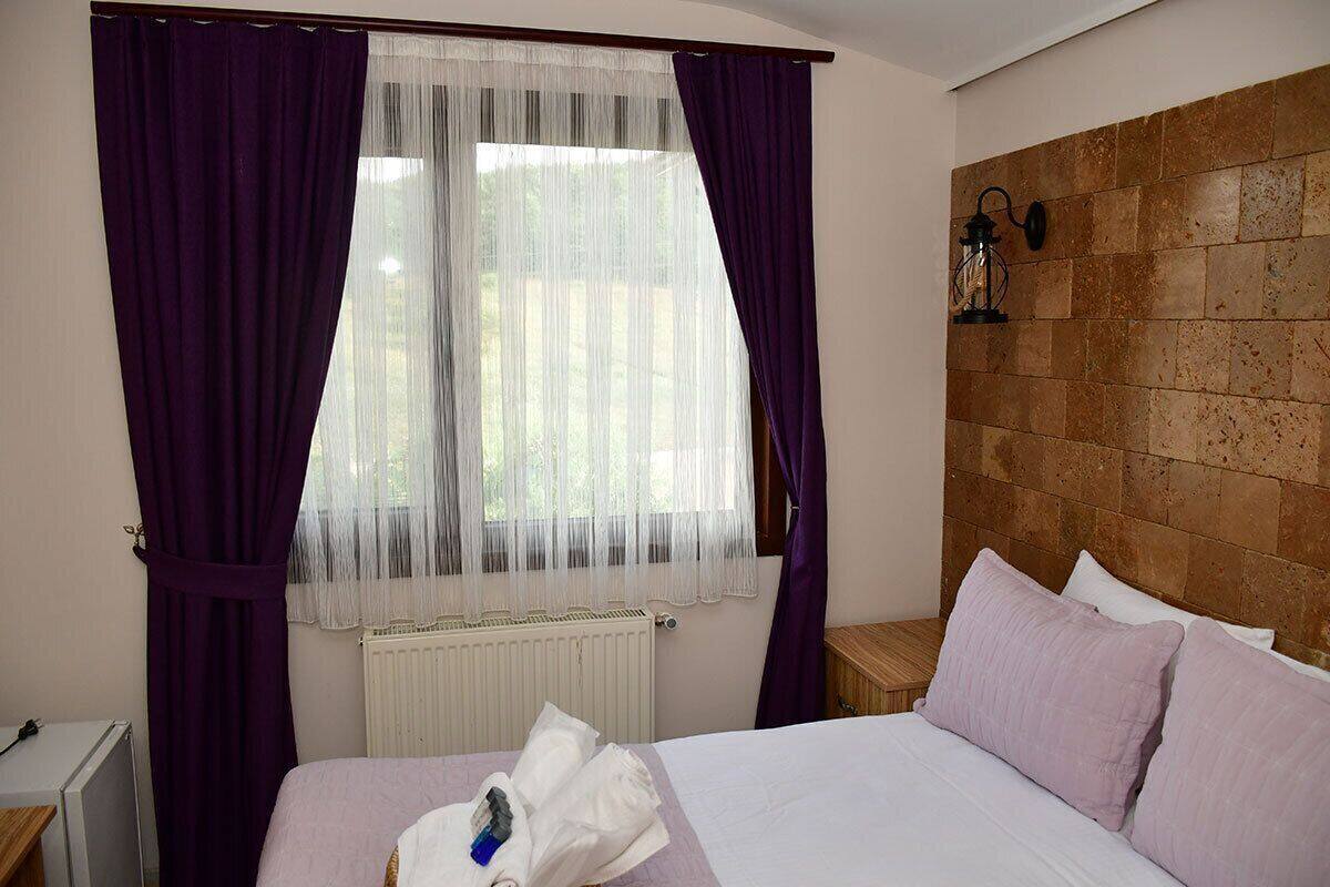 A hotel room with a bed, a window with purple curtains, a radiator, a wooden headboard, and a nightstand with a lamp.