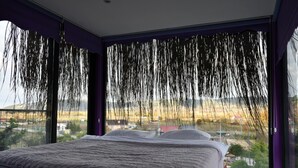 A bedroom with a large bed, a view of a rural landscape through the window, and purple curtains.