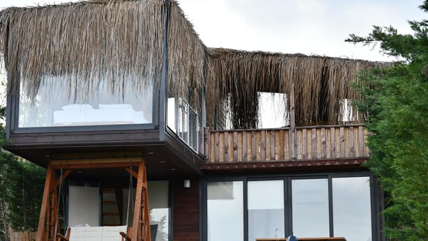 A two-story building with a thatched roof over the upper level, a stone wall in front, and a wooden deck with a swing chair and picnic table.