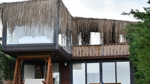 Un edificio de dos plantas con tejado de paja sobre el nivel superior, un muro de piedra en la parte delantera, y una terraza de madera con una silla mecedora y una mesa de picnic.