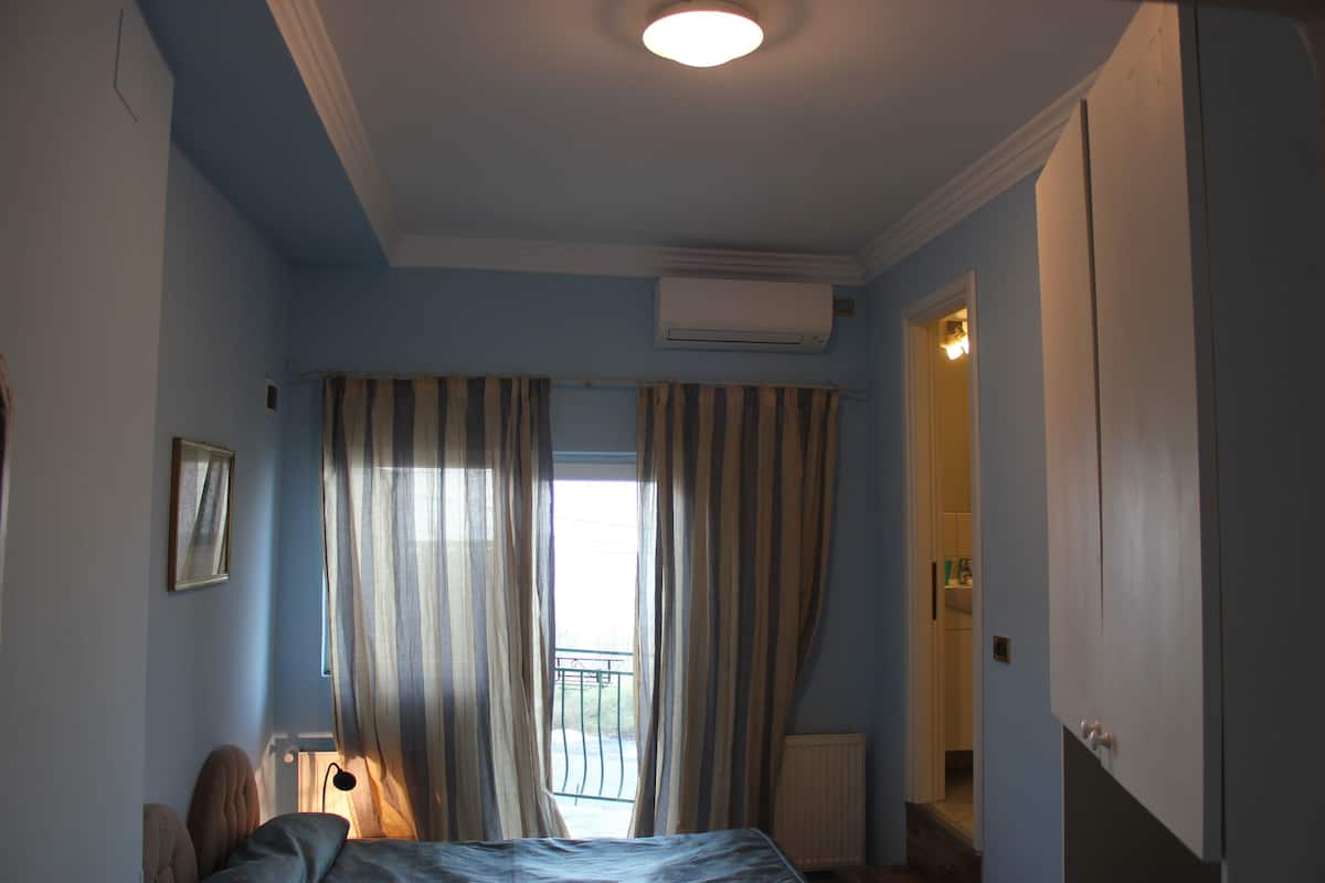 A bedroom with a bed, an air conditioning unit, a window with curtains, a bedside lamp, and a door leading to a bathroom.