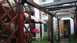 Una pérgola de madera con una mesa y sillas debajo, rodeada de follaje exuberante y flores rosadas.