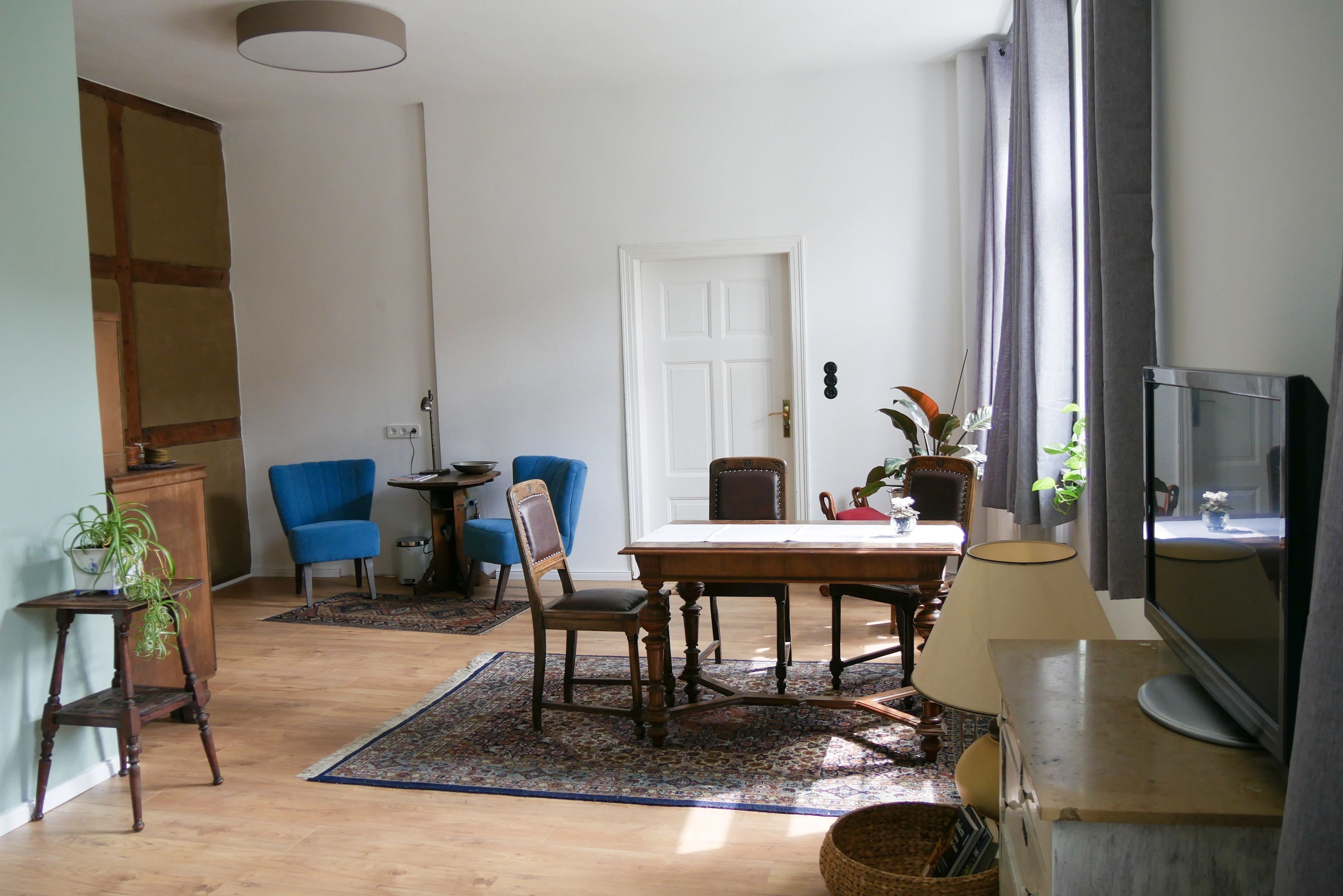 Una sala de estar con una mesa de comedor de madera, dos sillas azules, un televisor y un amplio ventanal con cortinas.