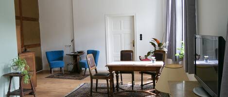 Una sala de estar con una mesa de comedor de madera, dos sillas azules, un televisor y un amplio ventanal con cortinas.