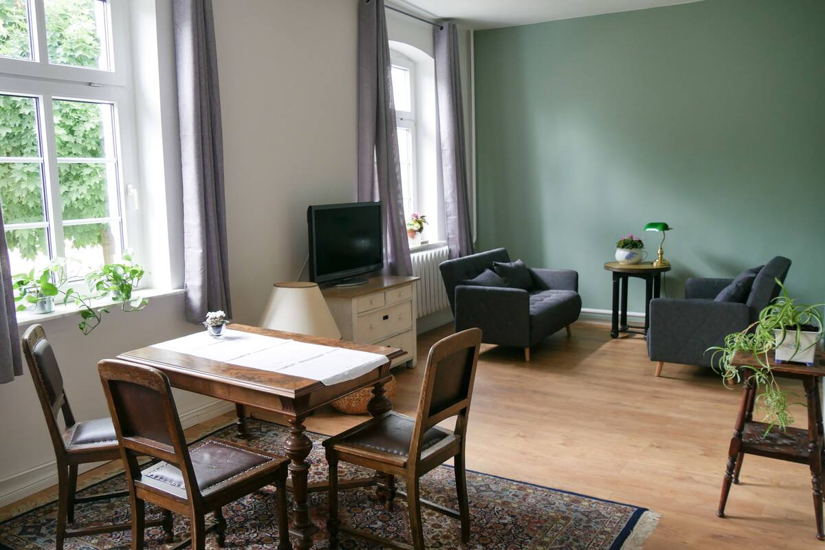 A living room with a dining table, chairs, a sofa, a television, and a small side table with a lamp and plant.