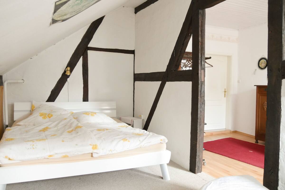 A bedroom with a white bed, wooden beams, a red rug, and a door leading to another room.