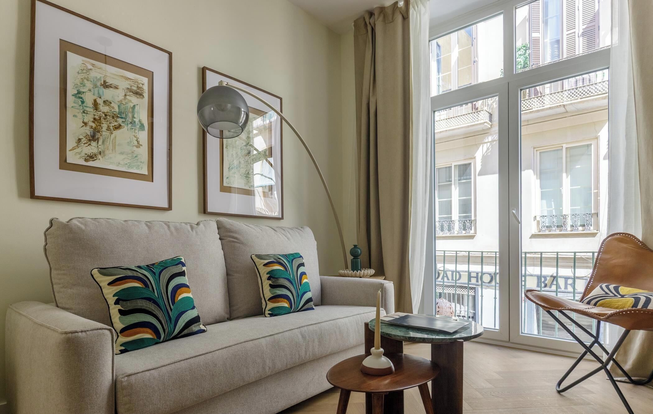 Sala de estar con sofá, almohadones coloridos, lámpara, mesita y silla. Se observan obras de arte enmarcadas en la pared y vistas de edificios a través de amplias ventanas.
