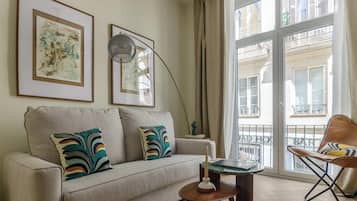 Sala de estar con sofá, almohadones coloridos, lámpara, mesita y silla. Se observan obras de arte enmarcadas en la pared y vistas de edificios a través de amplias ventanas.