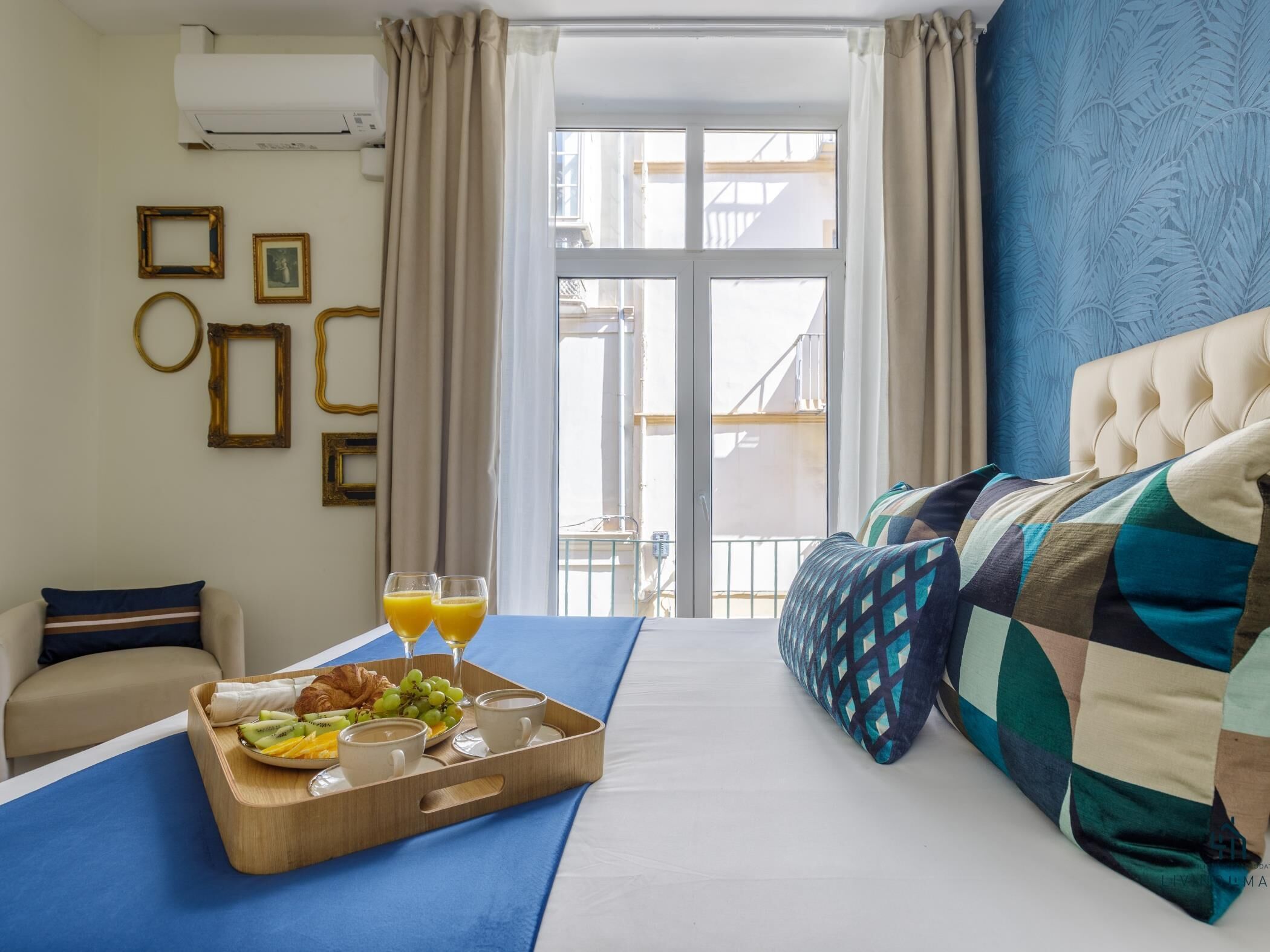 Habitación de hotel con cama, bandeja de desayuno con frutas y bebidas, sofá y vistas al exterior.