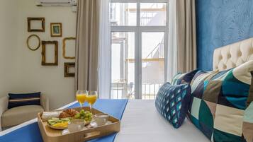 Habitación de hotel con cama, bandeja de desayuno con frutas y bebidas, sofá y vistas al exterior.