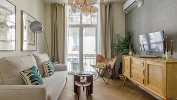 Un salón con sofá, mesa de centro, televisor, aparador y balcón con cortinas.