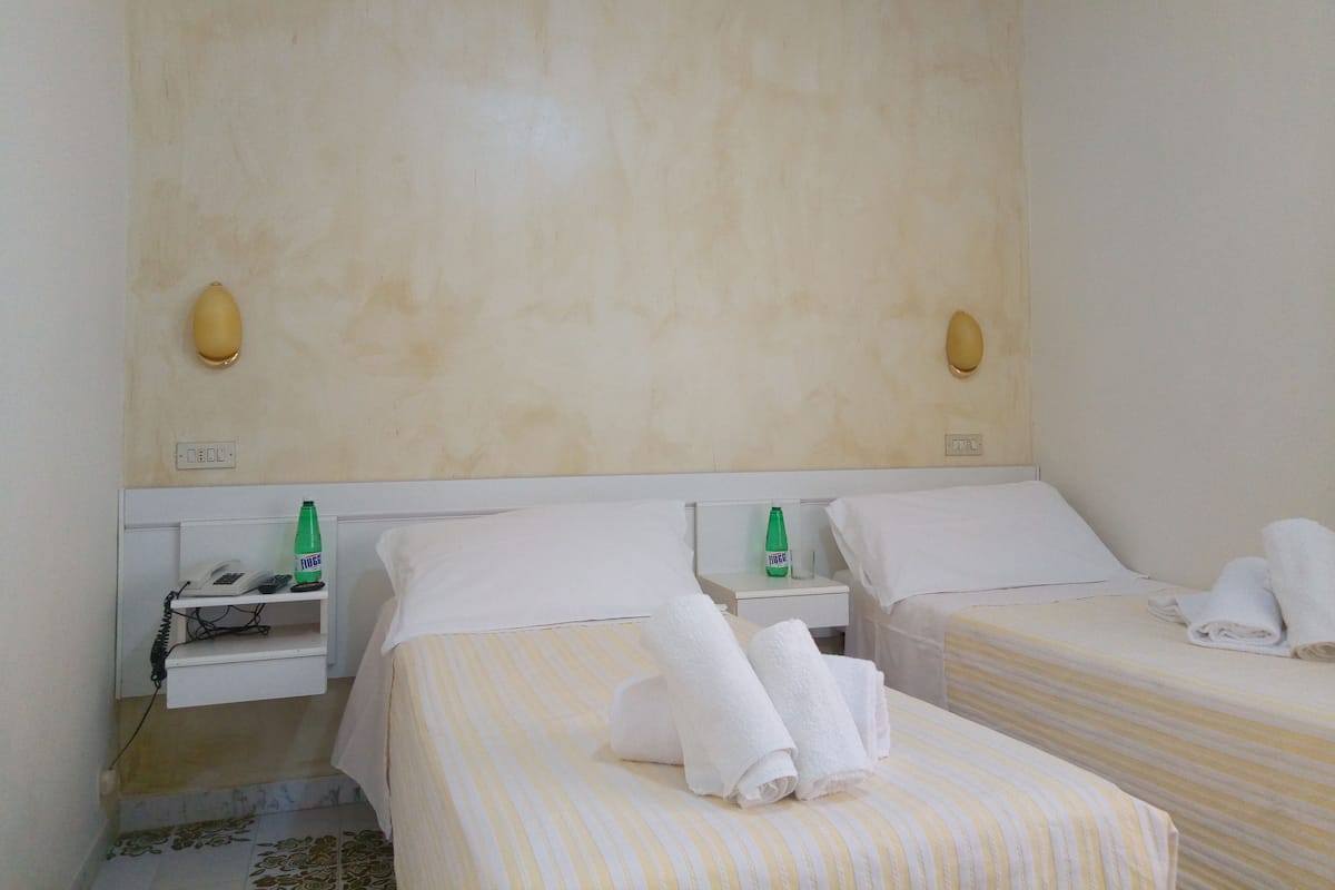 A hotel room with two beds, a bedside table with a phone and a bottle, and a wall-mounted light fixture.