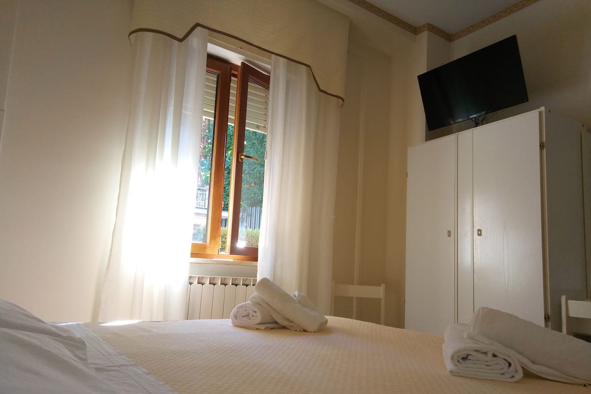 A hotel room with a bed, a window with curtains, a radiator, a television mounted on the wall, and a wardrobe.