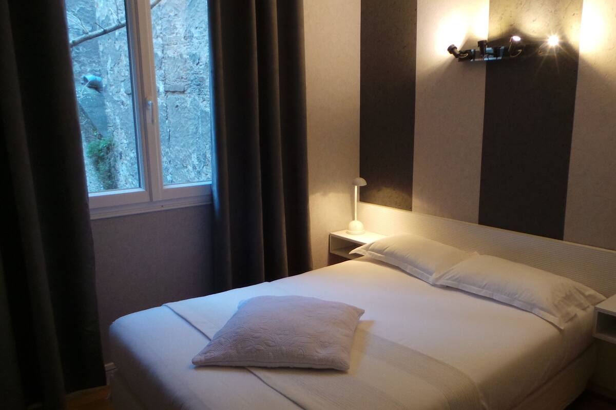 A neatly made bed with a white pillow and a folded white towel, a bedside table with a lamp, and a window with curtains.