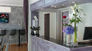 Un área de recepción con un diseño moderno, que incluye un mostrador con un florero, una pantalla de televisión y una zona de estar con sillas.