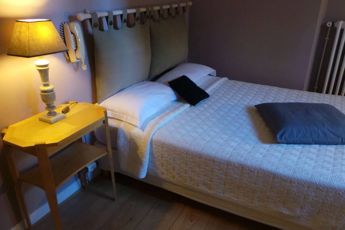 A neatly made bed with a quilted cover, a bedside table with a lamp, and a window with curtains.