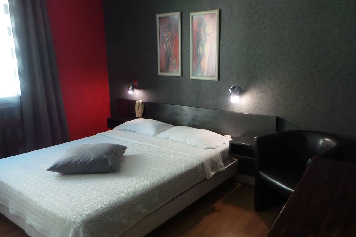 A hotel room with a bed, a chair, a desk, two bedside lamps, and two framed artworks on the wall.