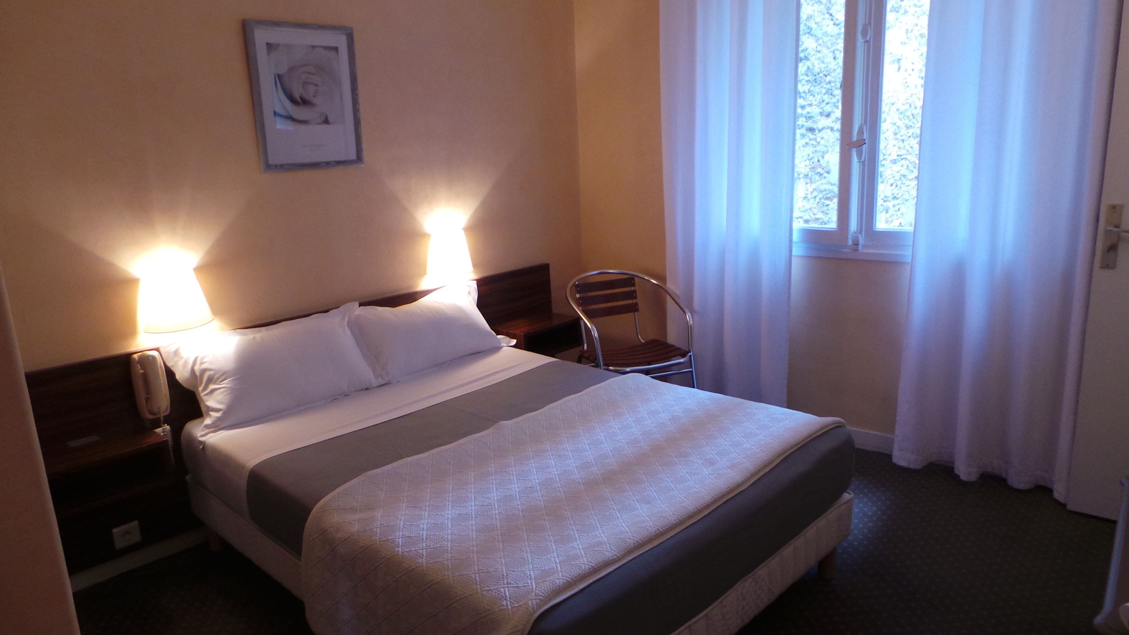 Une chambre d’hôtel avec un lit, une chaise, une table de chevet avec un téléphone et une fenêtre avec des rideaux.
