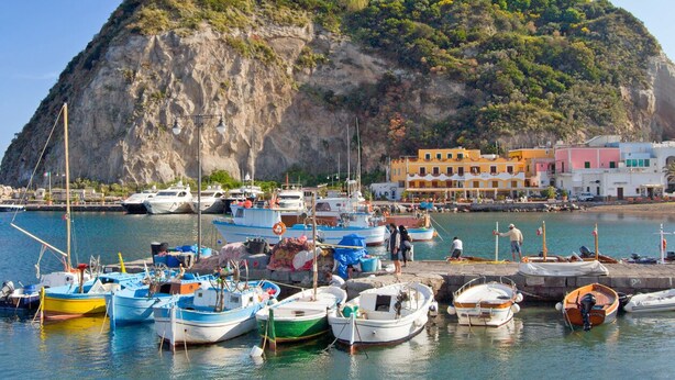 Un port où sont amarr és des bateaux, une colline rocheuse couverte de végétation et des bâtiments aux couleurs vives qui bordent le front de mer.