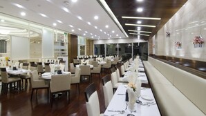 A spacious dining area with multiple tables and chairs, well-lit with ceiling lights, and a long bench along one wall. - Al Murooj Grand Hotel (Muscat)