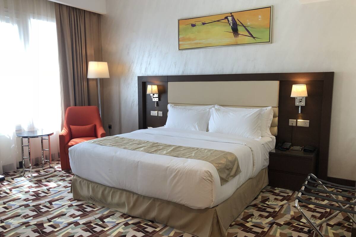A hotel room with a large bed, a chair, a desk, and a painting on the wall.