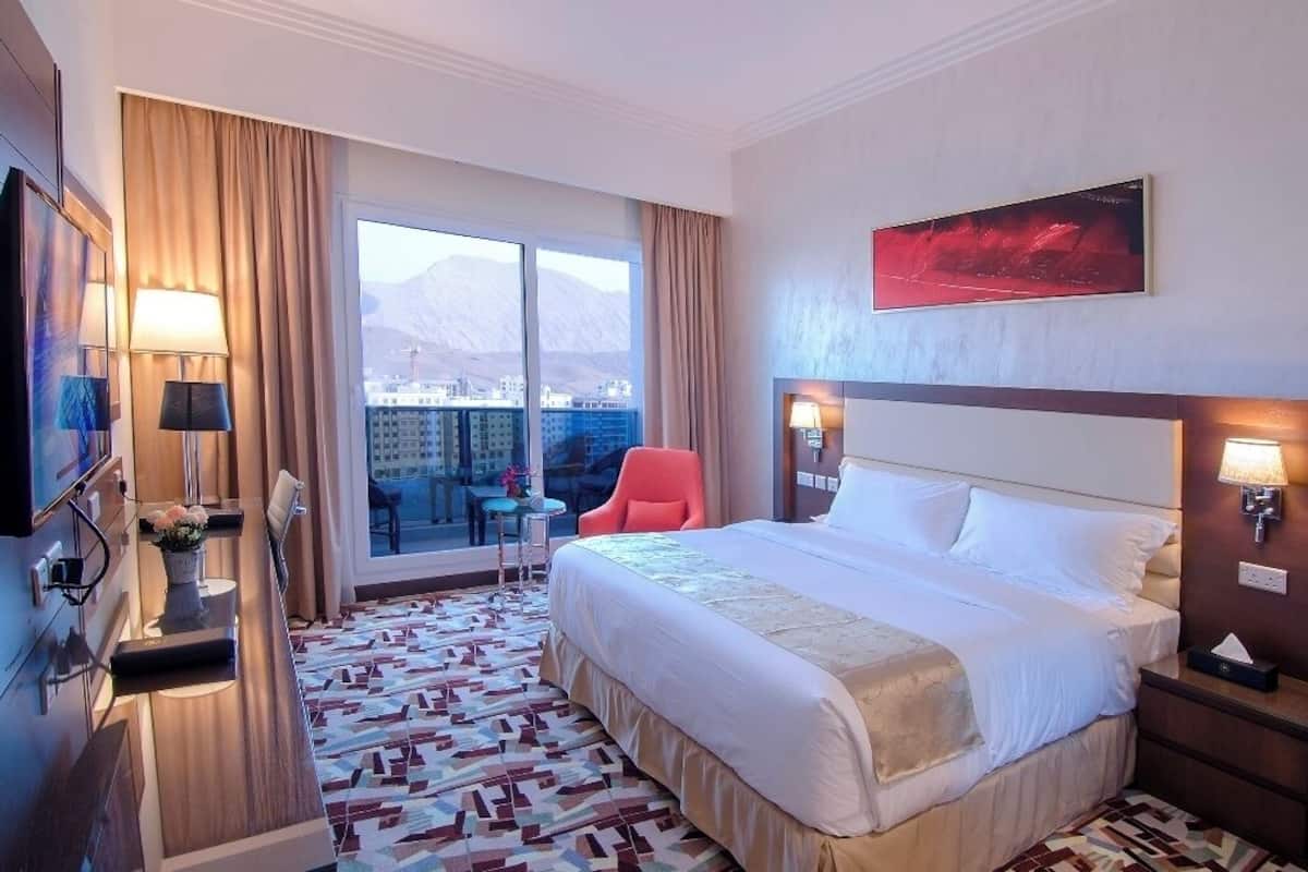 A hotel room with a large bed, a desk with a chair, a TV, a window with a view of a cityscape, and a painting on the wall.