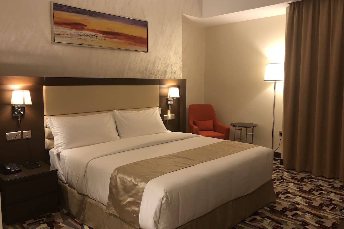 A hotel room with a large bed, a nightstand with a phone, a chair, a lamp, and a painting on the wall.