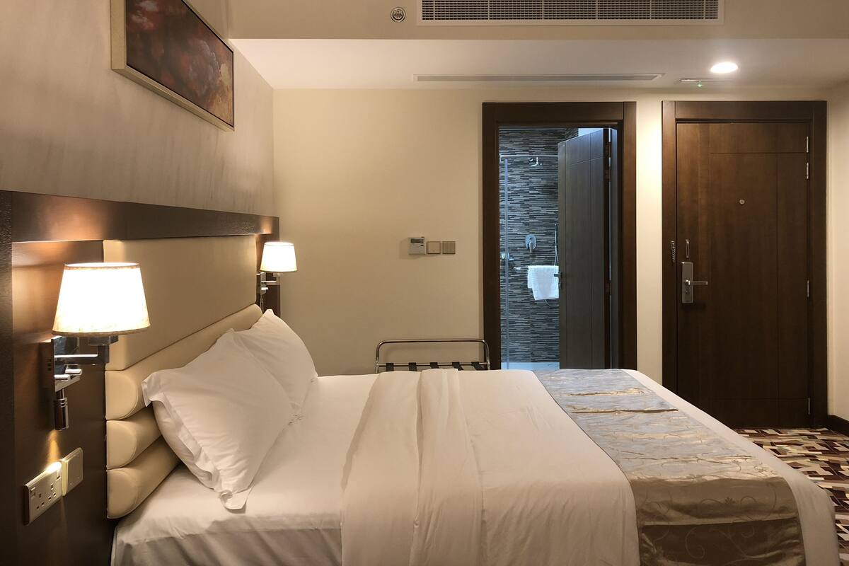 A hotel room with a bed, bedside lamps, a painting on the wall, a door, and a shower area.