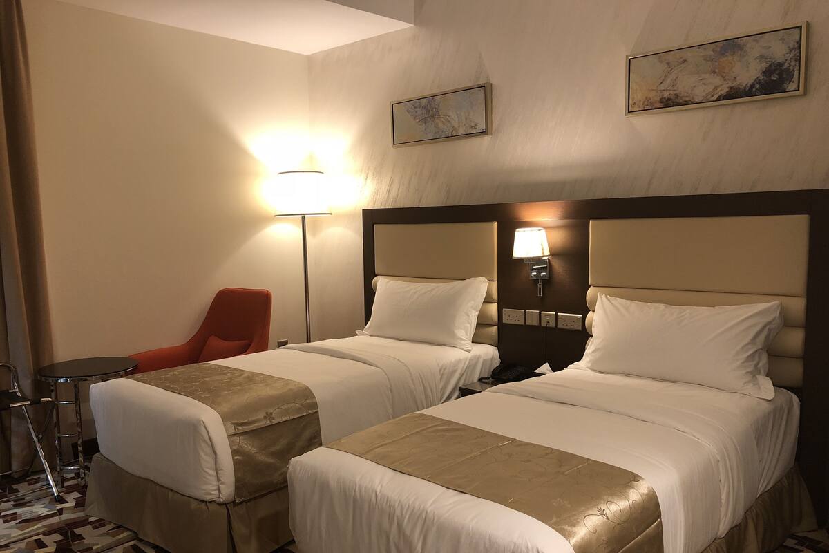 A hotel room with two beds, a red chair, a small table, and a floor lamp.