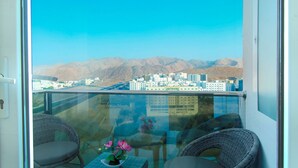 A balcony with wicker chairs and a small table with a flower vase, overlooking a cityscape with mountains in the background. - Al Murooj Grand Hotel (Muscat)